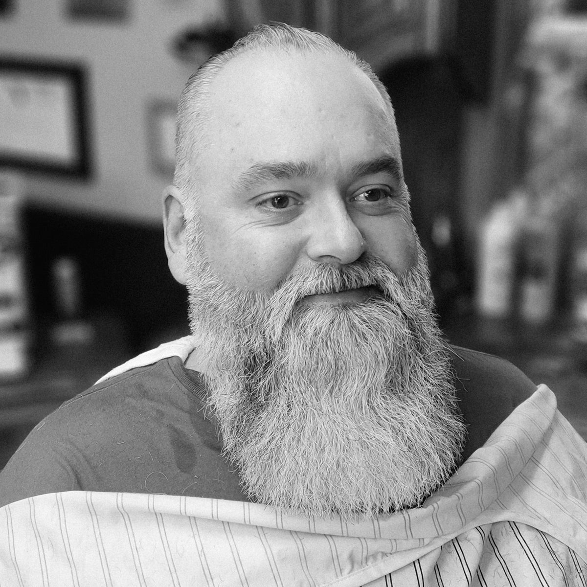 A black and white close-up portrait of a smiling man with a full gray beard, wearing a striped barber cape over a dark shirt in a barbershop setting.