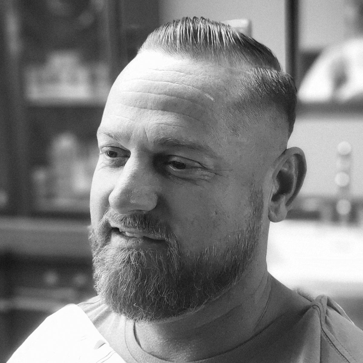 A black and white close-up portrait of a man with a short, styled haircut, fade, and beard, wearing a barber's cape in a barbershop setting.