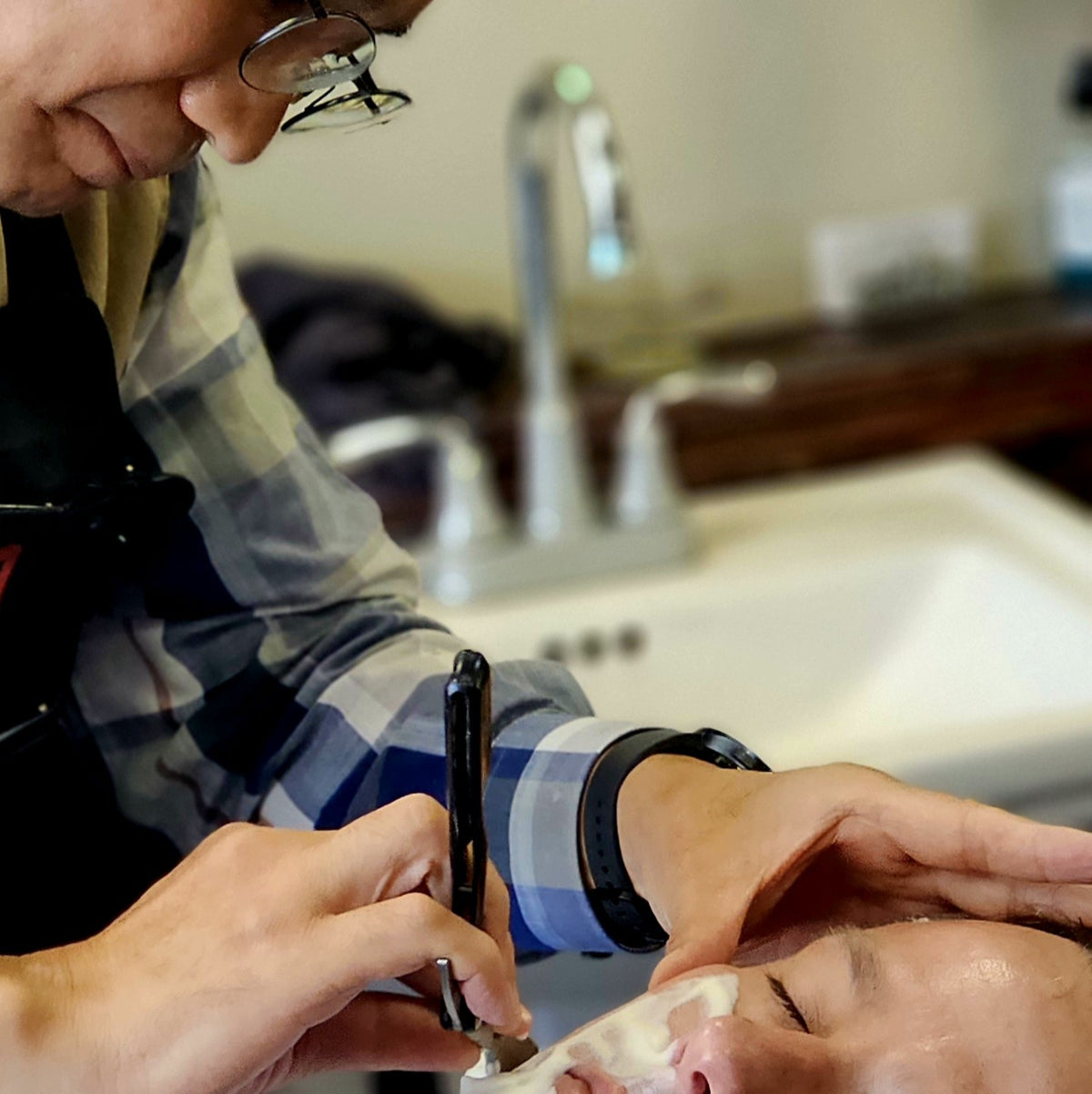 A close-up lifestyle shot of a barber shaving a client's face using a straight razor. The barber, wearing a blue and gray plaid shirt, is focused intently on the task, while the client lies under the barber's hands.