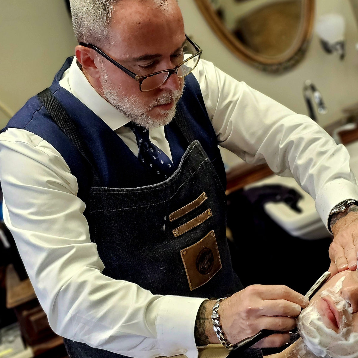 A man with a gray beard and glasses, wearing a white shirt, patterned tie, and dark denim apron, shaving another man's face in a barbershop setting.