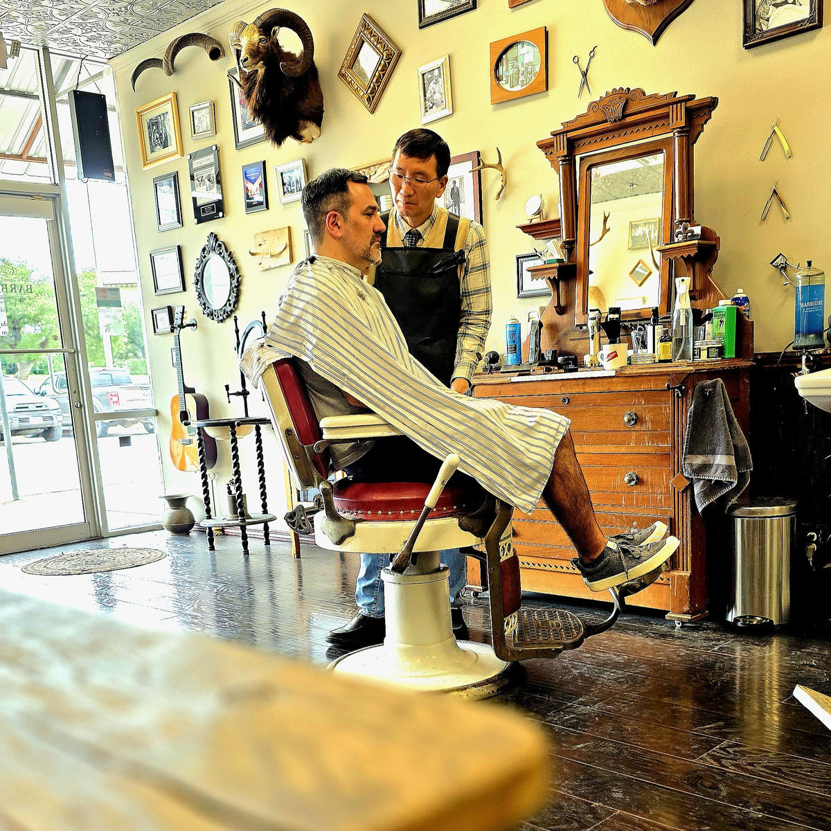 A man in a striped barber cape sits in a vintage red and cream barber chair while a barber in a black apron stands beside him in a detailed, rustic barbershop with wooden furniture and wall decor.