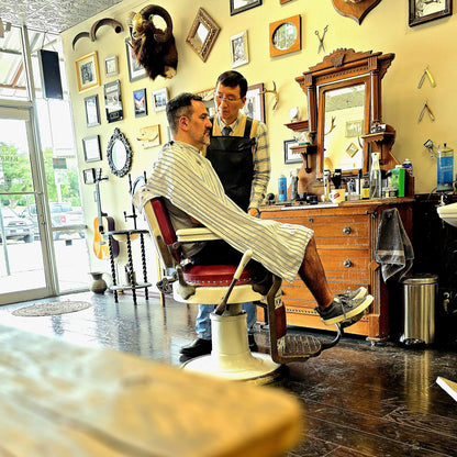 A man in a striped barber cape sits in a vintage red and cream barber chair while a barber in a black apron stands beside him in a detailed, rustic barbershop with wooden furniture and wall decor.