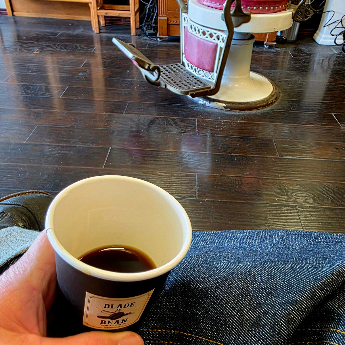 A hand holds a paper cup labeled 'BLADE BEAN' filled with dark coffee in a barbershop setting with dark wood floors and a vintage barber chair in the background.