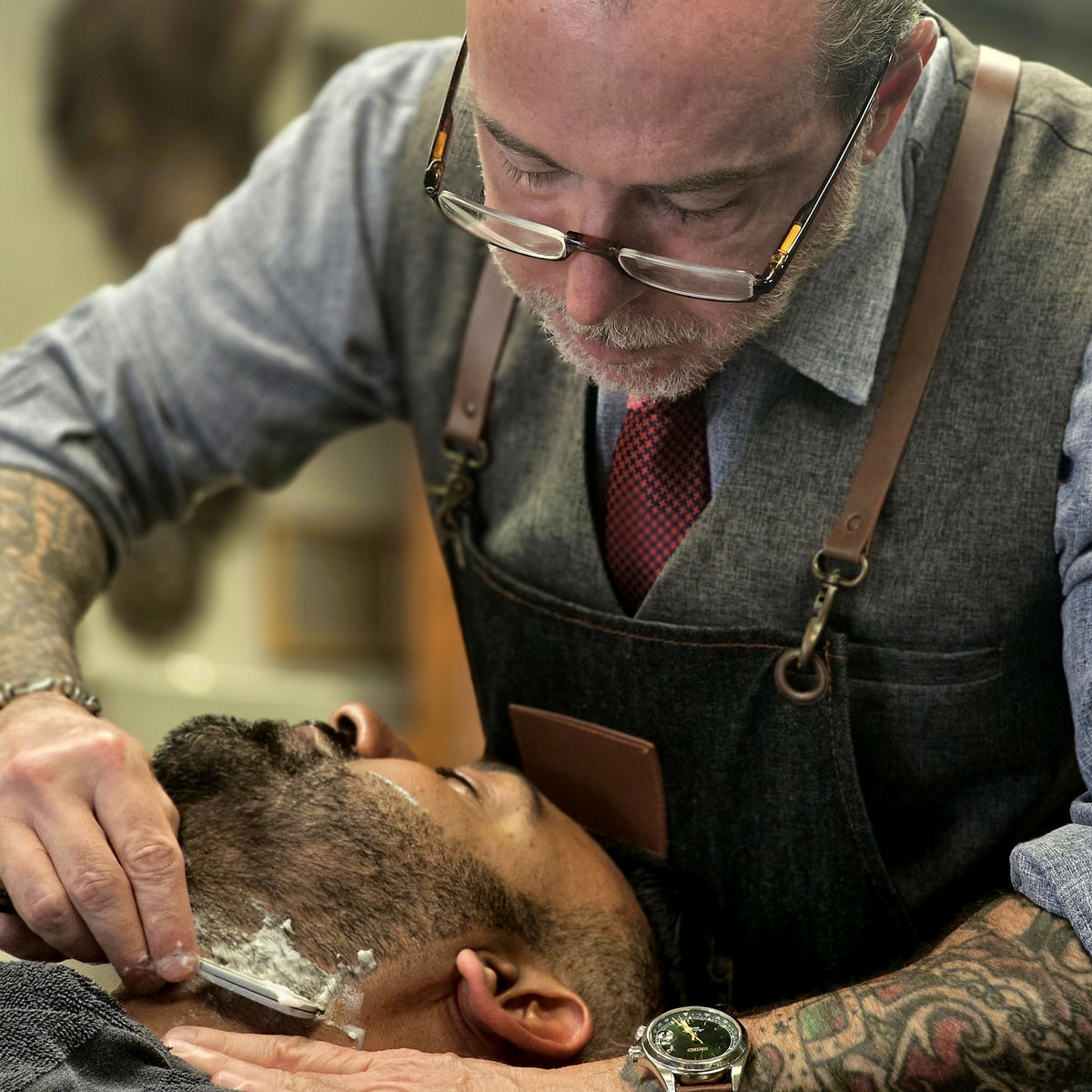 A close-up lifestyle shot of a professional barber with tattoos and glasses shaving a client's beard in a barbershop setting.