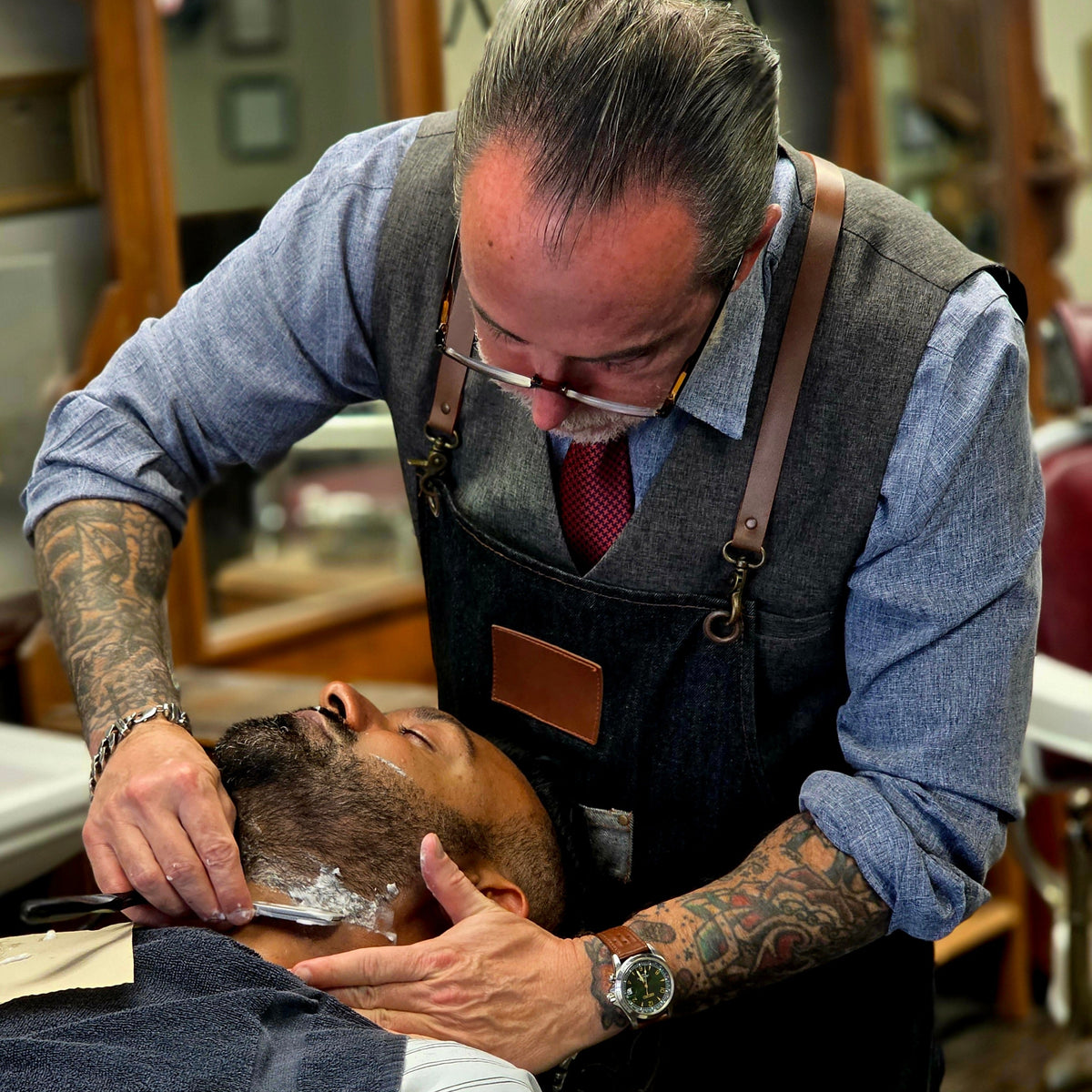 A professional barber with tattoos and glasses shaving a client's head in a barbershop. The barber is wearing a blue shirt, red tie, and dark gray apron with brown leather straps.