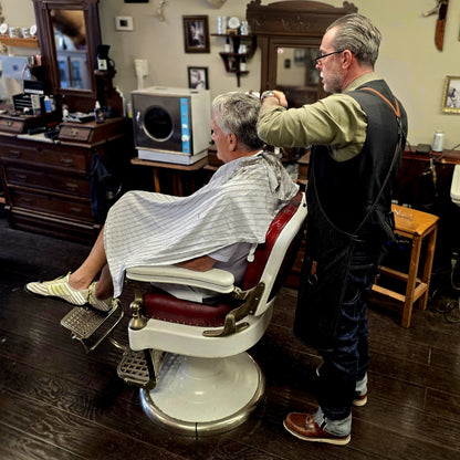A barber in a black apron cuts the hair of a customer sitting in a vintage-style barber chair inside a barbershop.