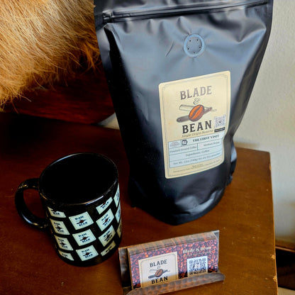 A black stand-up pouch of Blade Bean coffee sits on a wooden table next to a black ceramic mug with a white geometric pattern and a small branded card.