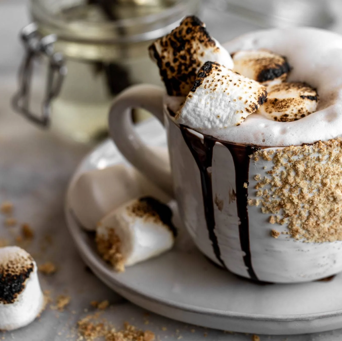 CAMPFIRE CUT coffee with marshmallows and chocolate drizzle in a white mug on a light surface.