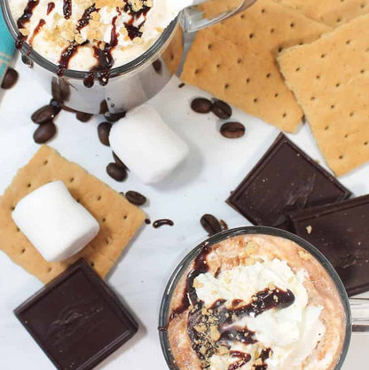 Two cups of CAMPFIRE CUT coffee with whipped cream, chocolate drizzle, and graham crackers on a white surface.