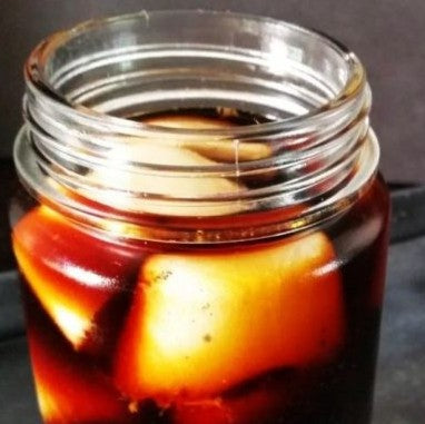 A clear glass mason jar filled with a dark amber liquid and large ice cubes, photographed close up against a dark background.