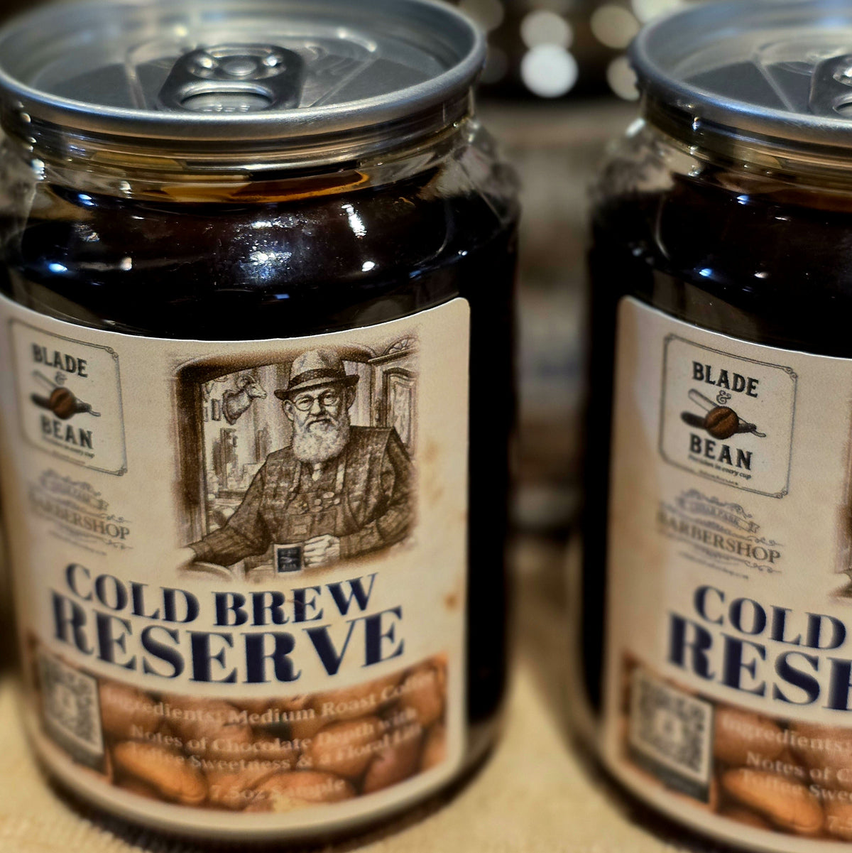 Two glass jars of Blade Bean Cold Brew Reserve coffee beans, with the front jar fully visible and the second jar partially cropped on the right, set against a warm, softly blurred background.