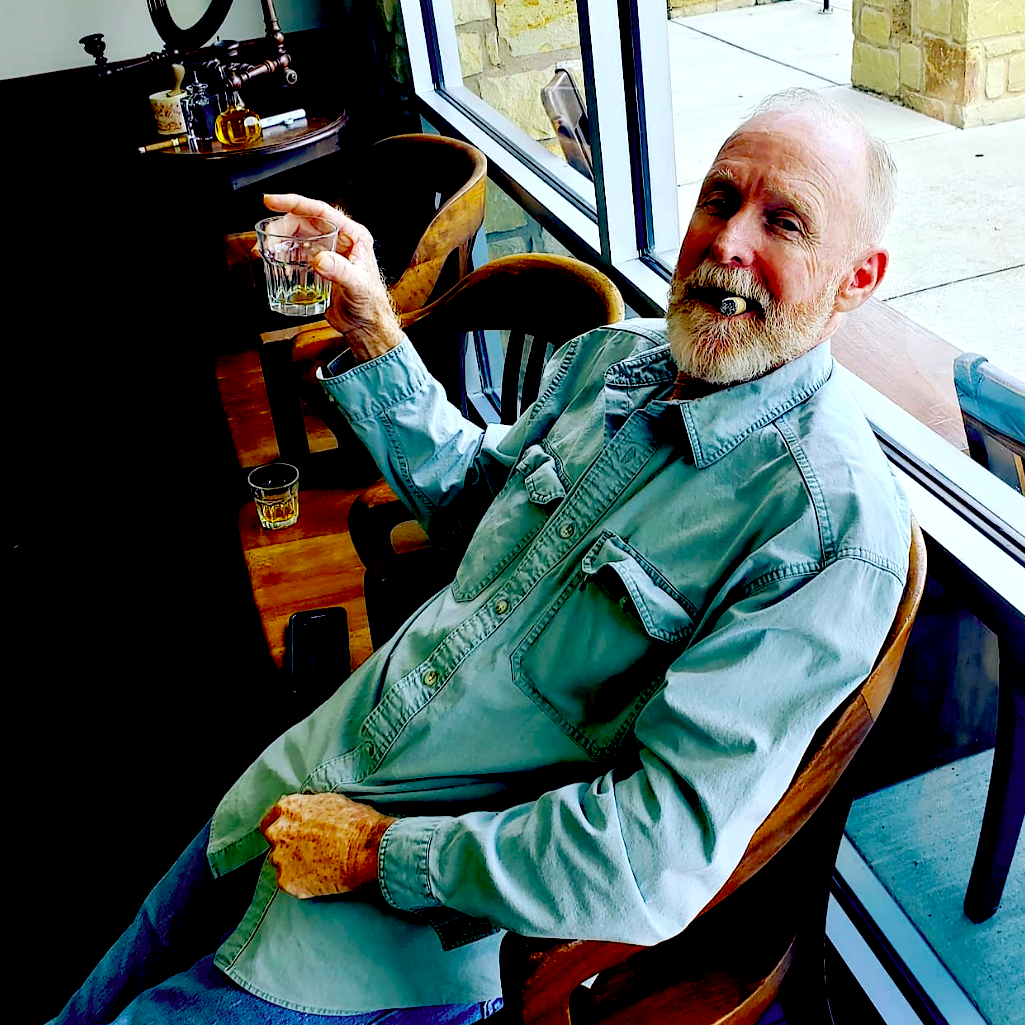 David Patrick sitting in Cedar Park Barbershop holding a glass of Bulleit bourbon, and smoking a Cuban cigar.