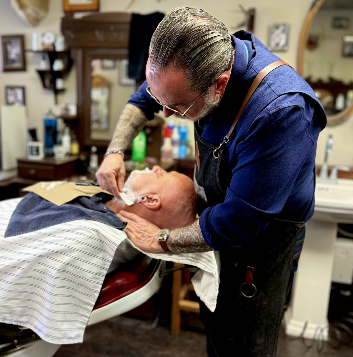 A professional barber with tattoos and glasses shaving a client's face with a razor in a vintage-style barbershop. The client is wearing a white and navy striped barber cape.