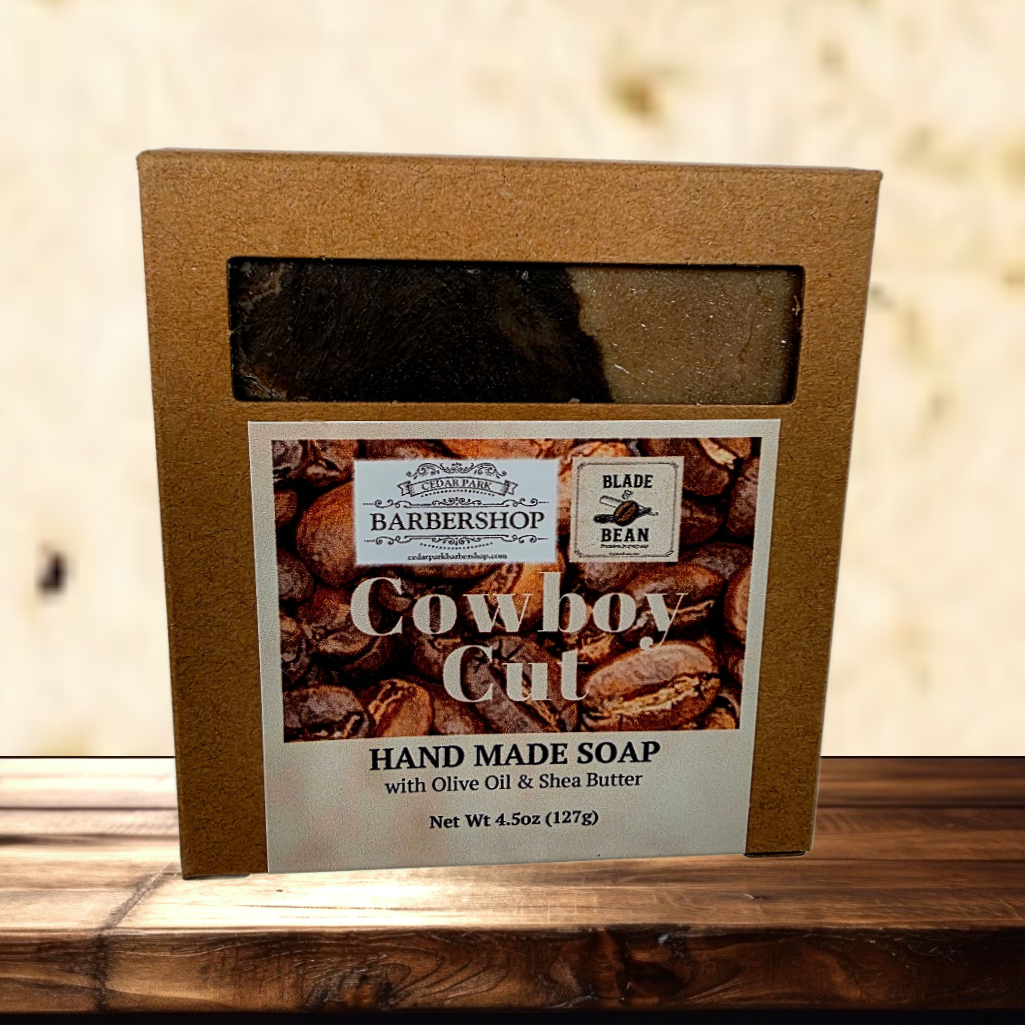 A boxed handmade soap product labeled 'Cowboy Cut' by Blade Barbershop, shown in a brown kraft box with a clear window and product label, placed on a wooden surface against a light textured wall.