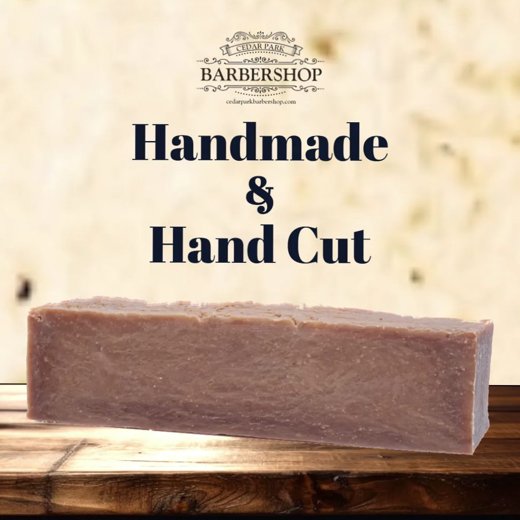 A rectangular bar of handmade soap in a dusty mauve-purple color, resting on a rustic wooden surface. The background is a textured beige wall with dark navy text reading 'Handmade & Hand Cut' and a barbershop logo at the top.