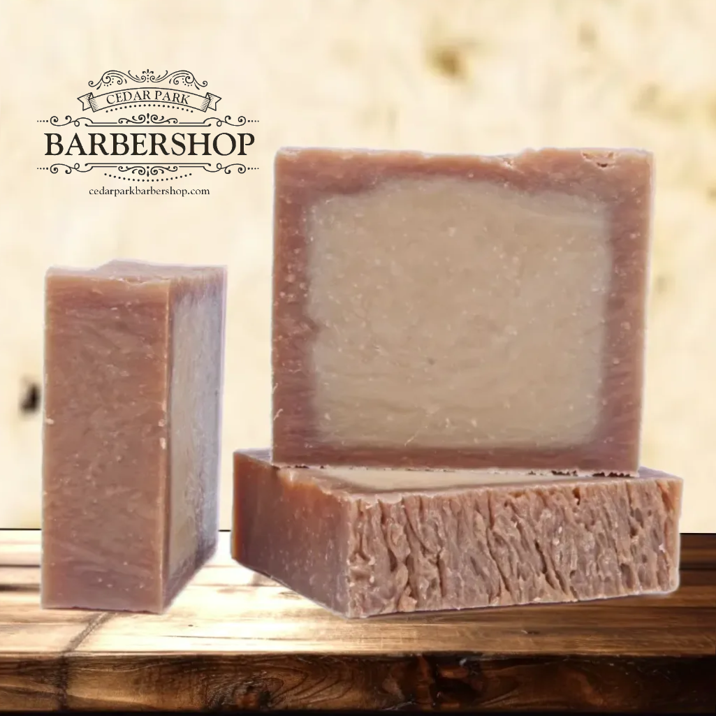 Three rectangular bars of lavender-colored soap displayed on a rustic wooden surface against a warm beige background. A brand logo for 'Cedar Park Barbershop' is visible in the upper left corner.