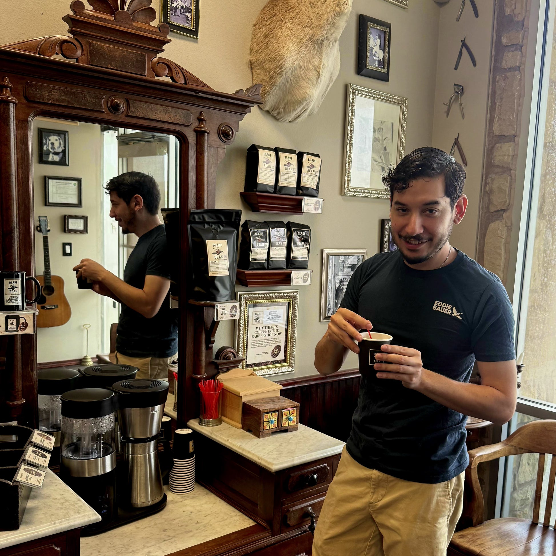 A man in a black t-shirt and khaki pants stands in a rustic coffee shop holding a paper cup, with a large wooden mirror behind him reflecting his image and shelves of coffee bags on the wall.