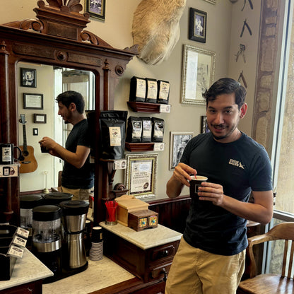 A man in a black t-shirt and khaki pants stands in a rustic coffee shop holding a paper cup, with a large wooden mirror behind him reflecting his image and shelves of coffee bags on the wall.