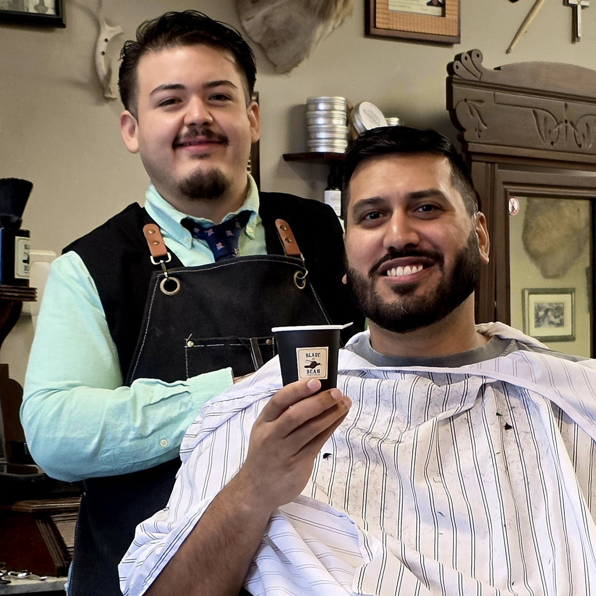 A smiling man in a barber's chair wearing a white striped barber cape holds up a small black cup with a logo, while another man in a black apron stands behind him in a vintage-style barbershop.