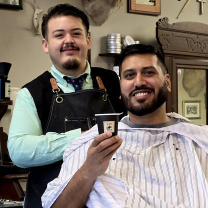 A smiling man in a barber's chair wearing a white striped barber cape holds up a small black cup with a logo, while another man in a black apron stands behind him in a vintage-style barbershop.