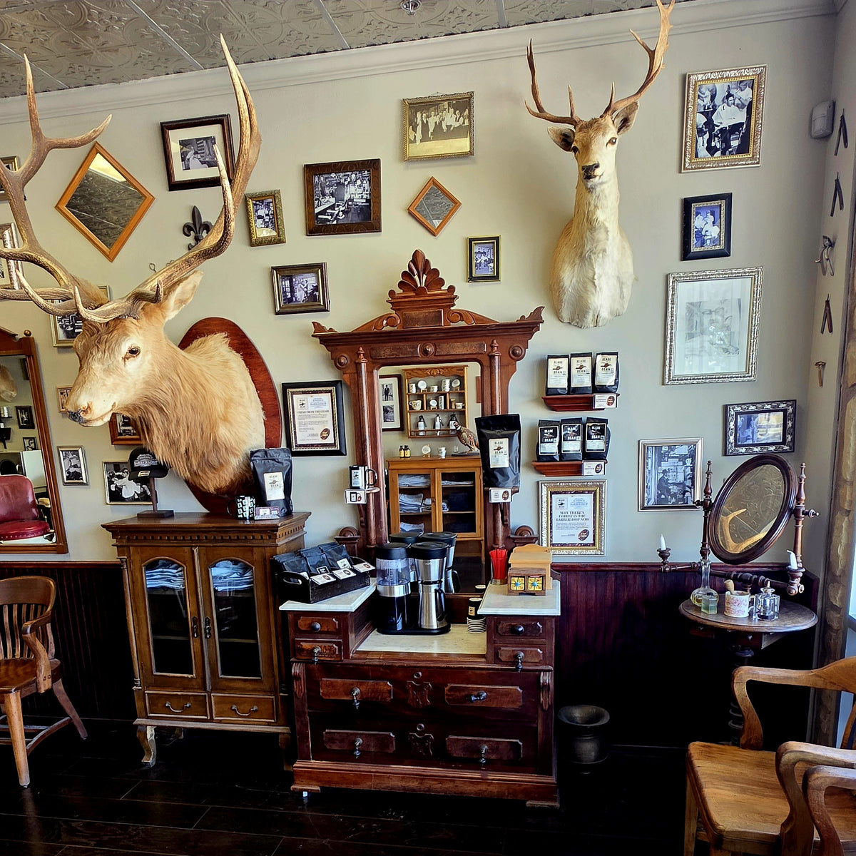 A rustic, vintage-style coffee station inside Cedar Park Barbershop, featuring two mounted deer heads with large antlers, framed black-and-white photographs, and antique wooden cabinetry. Coffee bags and mugs are visible on the counter.
