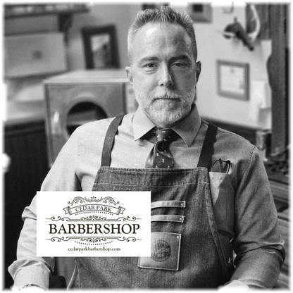 A black and white portrait of a man wearing a denim apron over a dress shirt and tie, standing in a barbershop environment. A white logo overlay at the bottom left reads 'CEDAR PARK BARBERSHOP' with 'cedarparkbarbershop.