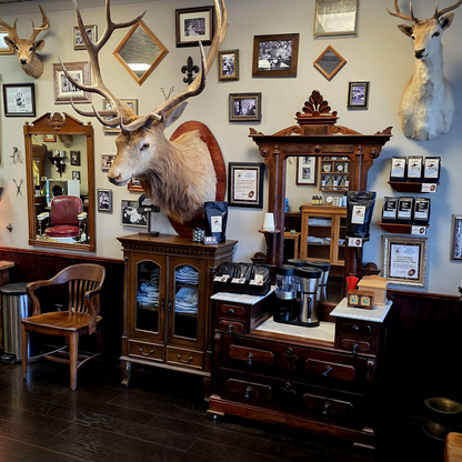 A rustic coffee bar area inside Cedar Park Barbershop, featuring a large mounted elk head with antlers on a wooden cabinet, additional taxidermy mounts on the wall, framed pictures, mirrors, shelves, and coffee equipment on counters.